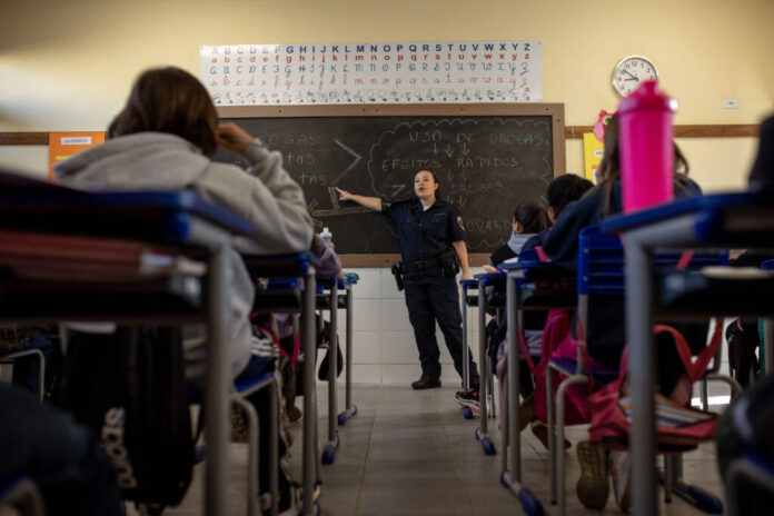 A imagem mostra uma sala de aula com uma policial fardada dando uma aula sobre o uso de drogas para um grupo de alunos. A policial está parada na frente de um quadro-negro onde escreveu "Uso de Drogas" e "Efeitos Rápidos". Ela está apontando para o quadro-negro enquanto fala para os alunos. Os alunos estão sentados em suas carteiras, e parecem estar prestando atenção à aula. A sala de aula é simples, com paredes brancas e carteiras azuis.